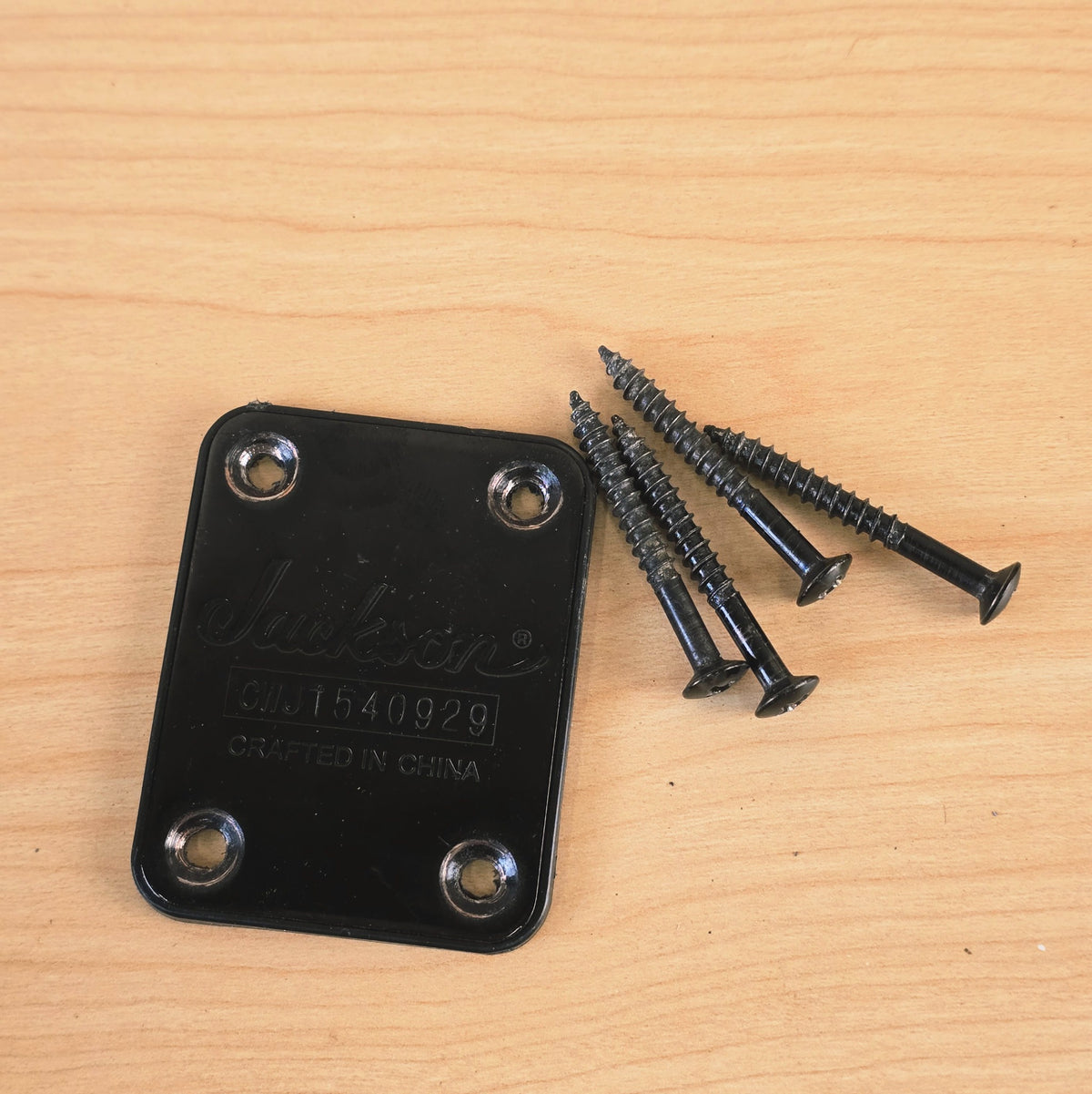 Jackson neck plate displayed alongside four black mounting screws on wooden surface