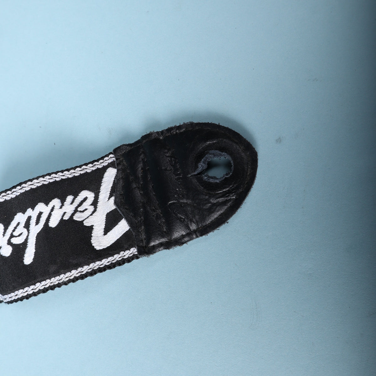 Close-up of the black leather end tab on a Fender running logo guitar strap showing strap button hole wear