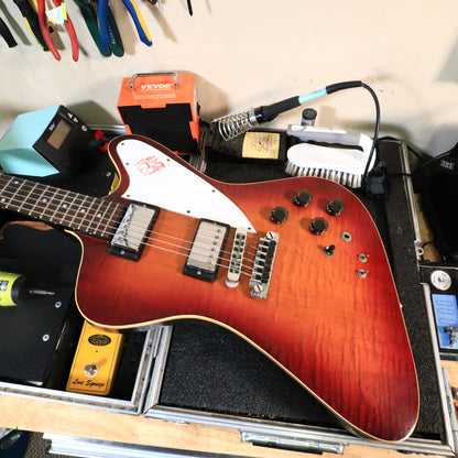 Front of the guitar resting on a workbench displaying the electronics layout and flame maple figuring.