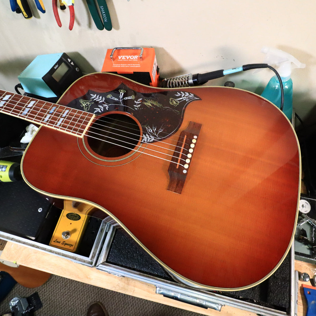 Gibson Early '60s Style Hummingbird acoustic guitar on a workbench showing the cherry sunburst top and ornate pickguard.