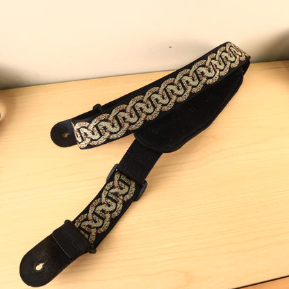 Planet Waves Celtic pattern guitar strap with black padded bass shoulder pad laid flat on a wooden surface.