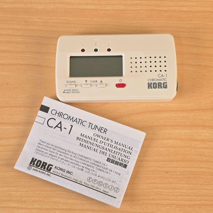 Top-down view of Korg CA-1 chromatic tuner next to its owner's manual on a wooden table.
