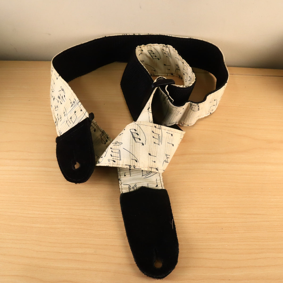 Cream sheet music print guitar strap coiled on a wooden surface showing black suede end tab.