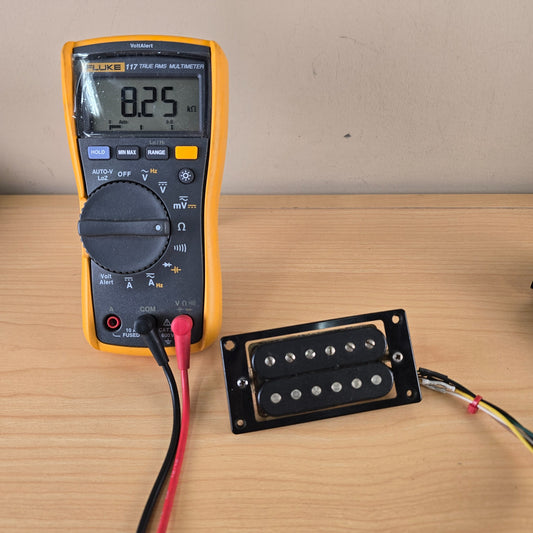Front view of black Schaller humbucker sitting alongside a Fluke multimeter measuring 8.25 kilo-ohms