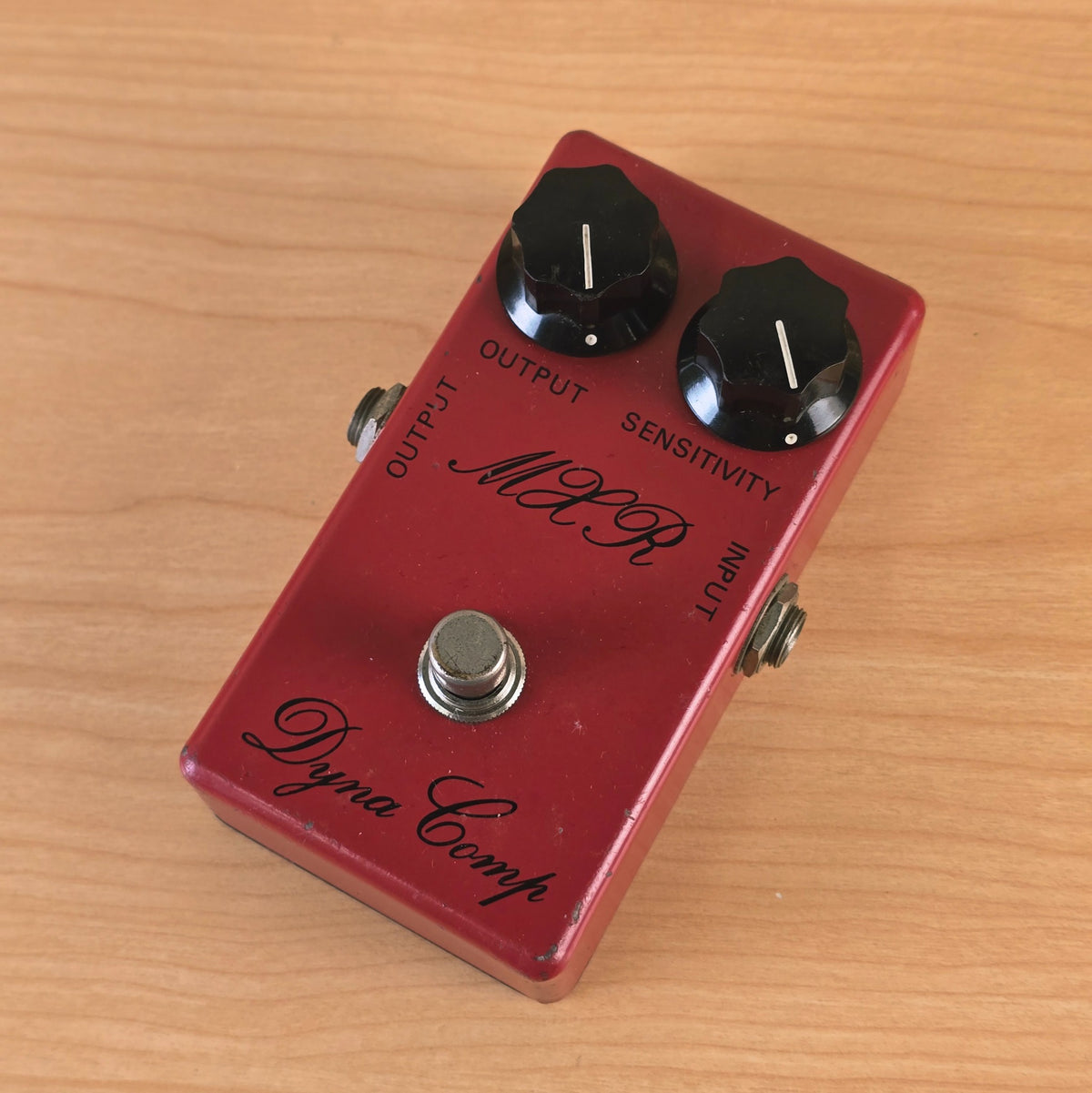 Three-quarter top view of the script logo MXR Dyna Comp pedal on a wooden surface.